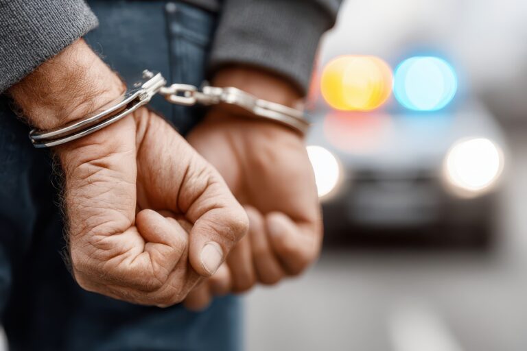 Close-up on hands cuffed with police car lights blurred in background. Concept of arrest, crime, and law enforcement, justice, and being apprehended by the police. Manos esposadas de un hombre detenido por la policía.