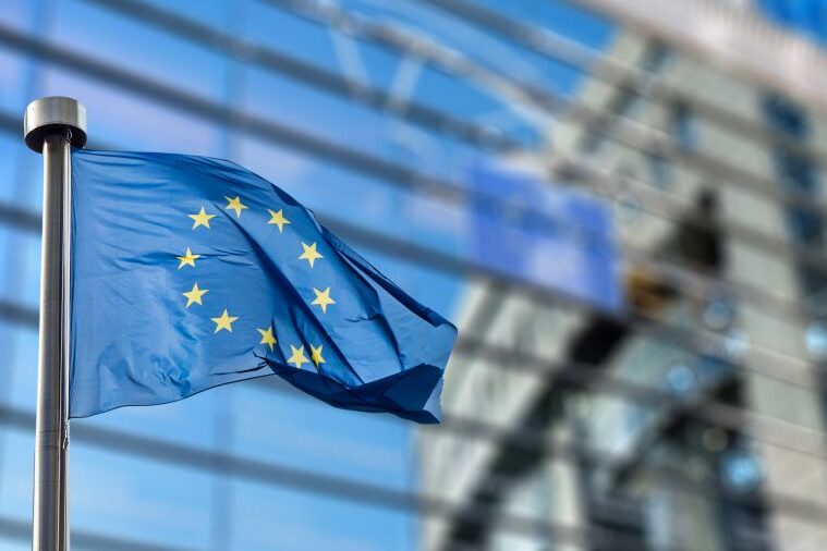 Union-Europea-Bandera-ok Bandera de la Unión Europea ondeando con el edificio del parlamento Europeo al fondo en Bruselas.