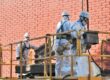 Trabajadores con trajes de protección contra sustancias peligrosas en amianto están encima de una plataforma y manipulan placas de fibrocemento.
