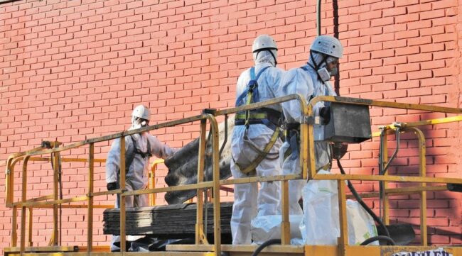 Trabajadores con trajes de protección contra sustancias peligrosas en amianto están encima de una plataforma y manipulan placas de fibrocemento.