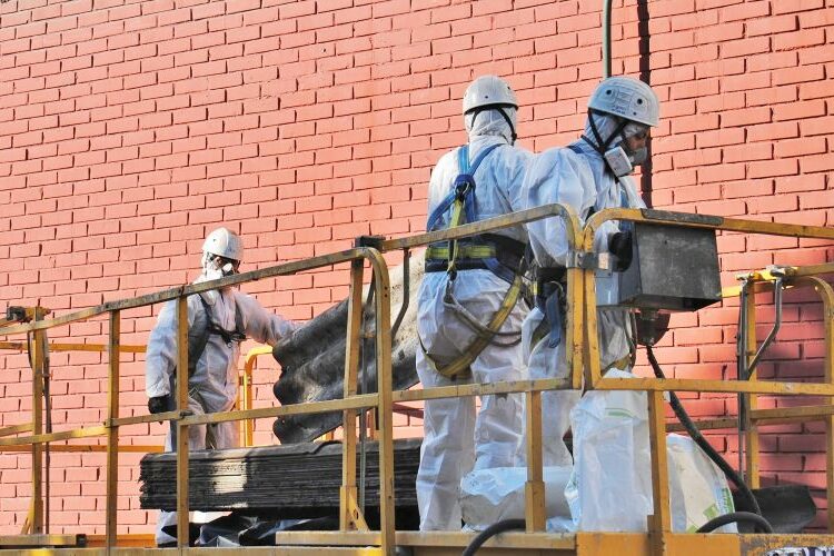 Retirada-amianto-asbesto-españa Trabajadores con trajes de protección contra sustancias peligrosas en amianto están encima de una plataforma y manipulan placas de fibrocemento.