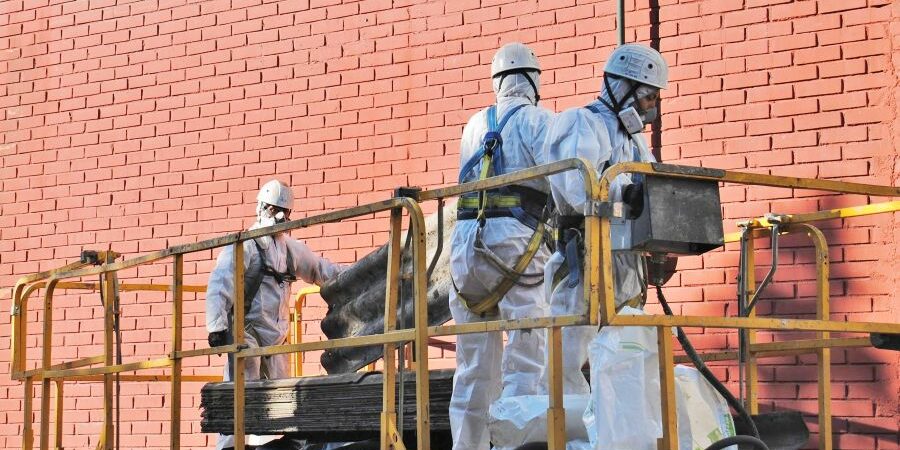 Trabajadores con trajes de protección contra sustancias peligrosas en amianto están encima de una plataforma y manipulan placas de fibrocemento.